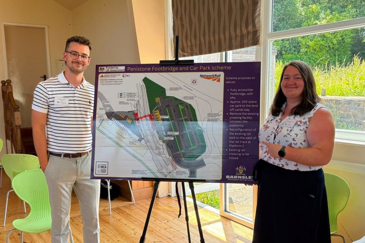 A photo of Alannah and Thomas standing in a community hall with the Penistone station image board of proposal in the middle